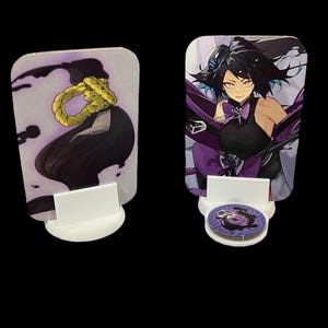 May include: Two rectangular anime-style character art cards, each mounted on a white stand. One card features a black and purple design with a yellow rope. The other card shows a woman with black hair and purple accents.