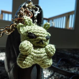 May include: A light green crocheted creature keychain featuring a small, textured body with pointed ears and black yarn embroidered eyes and mouth. The figure is attached to a silver-toned metal lobster clasp and chain, hanging from a dark bag.