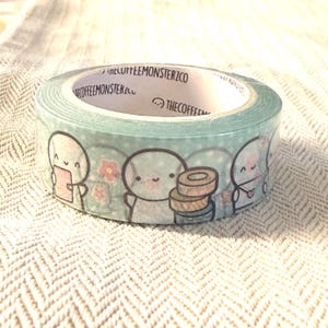 Puede incluir: Rollo de cinta washi decorativa con fondo azul claro, adornado con personajes de dibujos animados, flores y lunares. La cinta tiene el texto "THECOFFEEMONSTERCO" impreso en el interior. Ideal para manualidades.