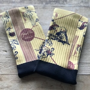 May include: A pair of yellow socks with a black cuff. The socks feature a vintage-style print of athletes playing different sports. The socks have the text "Pee Chee All Star Portfolio" printed on them.