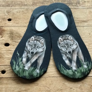 May include: A pair of black no-show socks featuring a wolf graphic. The wolf is grey and white and is walking towards the viewer. The socks are on a wooden surface.