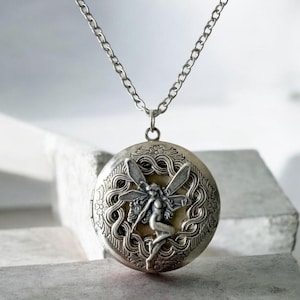 May include: A silver-toned circular locket pendant suspended from a matching link chain. The locket features an embossed relief of a winged fairy figure centered within an intricate, swirling filigree border with a matte, antiqued finish.
