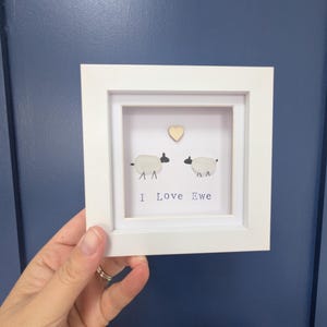 May include: A white framed picture with two sheep and a wooden heart. The text "I Love Ewe" is below the sheep. The artwork is held by a hand against a dark blue background.