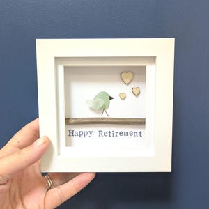 May include: A white framed artwork featuring a sea glass bird on a branch, with three wooden hearts. The text 'Happy Retirement' is printed below. The artwork is designed to be a retirement gift.