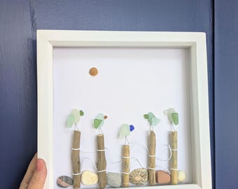 May include: A framed artwork featuring a whimsical beach scene. The art piece includes driftwood posts, sea glass birds, and pebbles, all set against a white background. A small wooden sun is placed at the top. The frame is white.