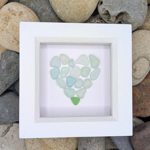 May include: A white framed artwork featuring a heart design made of sea glass. The sea glass pieces are various shades of blue, green, and white. The artwork is displayed on a bed of smooth, gray and brown stones.