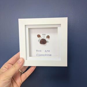 May include: A framed artwork featuring a crab made of small, brown stones. The crab is centered on a white background, with the words "You Are Clawesome" in blue below. The frame is white.
