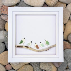 May include: A square white frame containing a piece of pebble art. Five small birds made of smooth, rounded stones in shades of green, white, and brown are perched on a piece of driftwood. The birds have thin wire legs and black beaks.