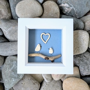 May include: A white framed artwork with a light blue backdrop. Two small, white bird figures sit on a wooden branch, with a wooden heart above. The artwork is set against a background of grey stones.