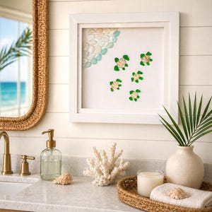 May include: A framed artwork depicting a beach scene with sea glass and turtle designs. The white frame surrounds a white background with a corner of white and blue sea glass and five green and shell turtles. The artwork hangs above a bathroom vanity.