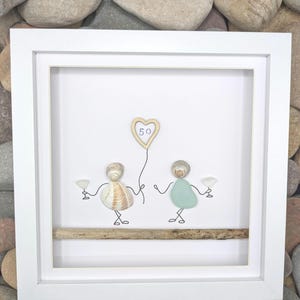 May include: A framed artwork depicting two figures made from seashells and sea glass, each holding a cocktail glass. A wooden heart-shaped balloon with the number 50 is suspended above. The artwork is set against a white background and framed in a white square frame.