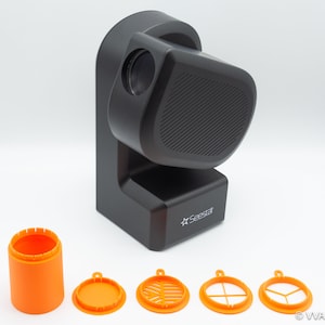 May include: A black projector with a speaker on the side and the word "Seestar" printed on the front. The projector is sitting on a white surface. There are four orange plastic discs with different designs on the surface.