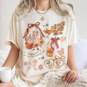 Può includere: T-shirt color avorio con una collezione di grafiche Disney a tema autunnale. Il design include un castello, zucche, Cip e Ciop, Topolino e Minnie e foglie autunnali. La maglietta ha maniche corte e una vestibilità ampia.