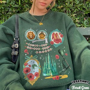 Puede incluir: Sudadera verde bosque con ilustraciones de El Mago de Oz. El diseño incluye imágenes del León, el Espantapájaros, los zapatos rojos y la Ciudad Esmeralda. También se ve el texto "SWEATSHIRT" y "Forest Green".