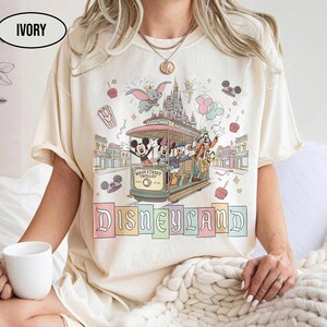 May include: Ivory-colored t-shirt featuring a colorful Disneyland graphic. The design includes Mickey Mouse, Donald Duck, Goofy, and other characters on a Main Street Trolley, with the Disneyland logo below. The shirt has short sleeves.
