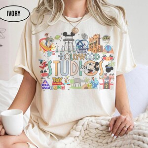Può includere: T-shirt color avorio con una grafica colorata che mostra le attrazioni e i personaggi di Hollywood Studios. Il design include il testo "Hollywood Studios", "Toy Story" e altre immagini relative al parco. La maglietta ha una vestibilità ampia.