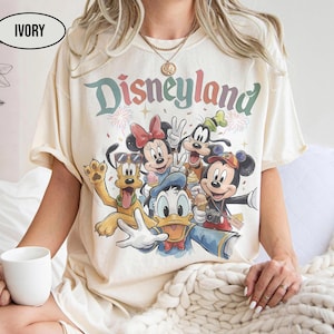 May include: Ivory-colored t-shirt featuring a colorful graphic of Disney characters, including Mickey Mouse, Minnie Mouse, Donald Duck, Goofy, and Pluto. The word "Disneyland" is printed above the characters in a rainbow font. The shirt has short sleeves.