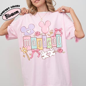 May include: A pale pink t-shirt with the word "magical" in colourful block letters. The shirt features cartoon graphics of a Mickey Mouse head, a balloon, a wand, a bunny, and roses. The text "Comfort Colors Blossom" is in a circle at the top.