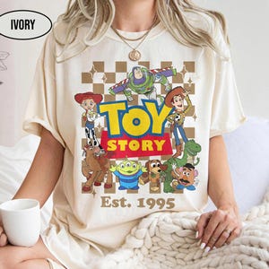 May include: Ivory-colored t-shirt featuring a graphic of Toy Story characters, including Woody, Buzz Lightyear, and others, with the words "TOY STORY" and "Est. 1995". The design is set against a brown and white checkered background.