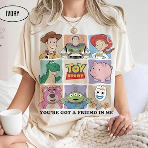 May include: Ivory-colored t-shirt featuring a colorful graphic of Toy Story characters. The design includes Woody, Buzz Lightyear, Jessie, Rex, Hamm, Lotso, the Aliens, and Forky. The text "TOY STORY" and "YOU'RE GOT A FRIEND IN ME" are also displayed.