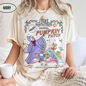 May include: Ivory-colored t-shirt featuring a whimsical illustration from a fairy tale. The design includes a fairy godmother, mice, a glass slipper, and the text "Fairy Godmothers Enchanted Pumpkin Patch." The shirt also has the text "Est. 1950" and "Bibbidi-Bobbidi-Boo!"