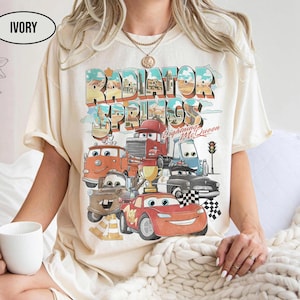 May include: An ivory t-shirt featuring a graphic print of Radiator Springs with various cartoon cars, including a red race car, a brown tow truck, and a gold trophy. The text reads Radiator Springs and Lightning McQueen in a retro font.