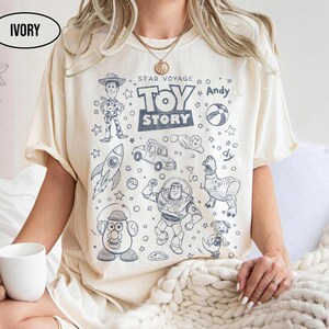 Puede incluir: Camiseta de color marfil con un diseño gráfico de personajes de la película animada "Toy Story". El diseño incluye a Woody, Buzz Lightyear y otros personajes, junto con el texto "Toy Story" y "Star Voyage + Andy".