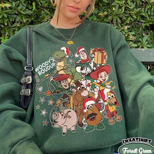 Op de afbeelding: Bostgroen sweatshirt met een afbeelding van Toy Story-personages met kerstmutsen. Het ontwerp bevat Woody, Buzz Lightyear en andere personages. De tekst "WOODY'S ROUDUP" staat links en "SWEATSHIRT Forest Green" rechtsonder.