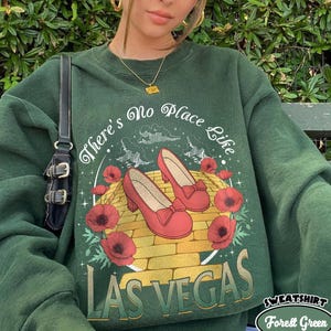 May include: Forest green sweatshirt with the text "There's No Place Like Las Vegas." The graphic features red shoes, poppies, and a yellow brick road design. The sweatshirt is a crew neck style.