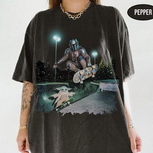 Pu&ograve; includere: Una t-shirt grigio antracite con una stampa grafica di un personaggio in armatura metallica che fa skateboard in una bowl, con una piccola creatura verde su uno skateboard sotto. Il testo PEPPER appare in un ovale nero in alto a destra.
