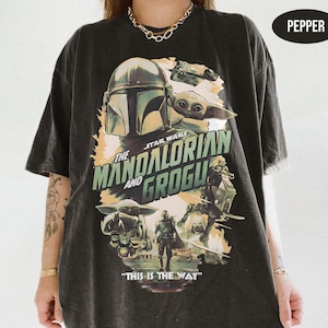 May include: Dark gray t-shirt featuring a graphic from the Star Wars series "The Mandalorian". The design includes the Mandalorian, Grogu, and the text "The Mandalorian and Grogu" and "This is the Way".