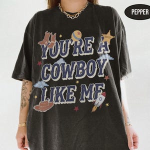 Puede incluir: Una camiseta de manga corta gris carb&oacute;n con el texto YOU'RE A COWBOY LIKE ME en letras azules. El gr&aacute;fico incluye ilustraciones de botas de vaquero marrones, una pelota, una estrella de sheriff, nubes, un sombrero marr&oacute;n, un cohete y estrellas rojas.