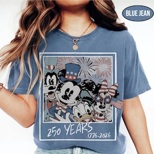 May include: A blue jean colored t-shirt featuring a graphic of cartoon characters wearing patriotic hats and accessories with fireworks in the background. The text 250 YEARS 1776-2026 is printed below the characters in a white, hand-drawn style font.