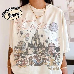May include: An ivory t-shirt featuring a graphic print of a castle, the moon, fireworks, and various cartoon characters. The text reads The Happiest Place On Earth and Walt Disney World. The shirt has short sleeves and a crew neckline.