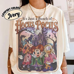 May include: An ivory t-shirt featuring a graphic print with the text "It's Just a Bunch of Hocus Pocus" above an illustration of three cartoon characters dressed as witches in front of a dark castle, bats, and a full moon in a framed design.