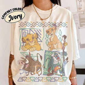 May include: An ivory t-shirt featuring a graphic print of characters from The Lion King, including Simba, Nala, Pumbaa, Timon, Rafiki, and Zazu, arranged in four rectangular panels with geometric borders and the text COMFORT COLORS IVORY.