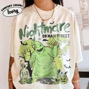 Puede incluir: Camiseta color marfil con un gr&aacute;fico verde que dice "Nightmare on Main Street". El dise&ntilde;o presenta un personaje verde, fantasmas, murci&eacute;lagos y calabazas. La camiseta tiene un corte holgado y mangas cortas.