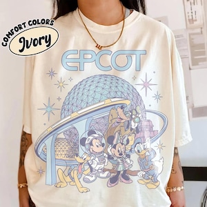 May include: An ivory t-shirt featuring a graphic print of the Epcot geodesic dome, a monorail, and cartoon characters including Mickey Mouse, Minnie Mouse, Pluto, Goofy, and Donald Duck. The text EPCOT is printed in large blue letters above.