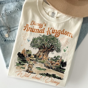 May include: Cream-colored t-shirt featuring a graphic of Disney's Animal Kingdom with Mickey Mouse. The shirt has the text "Disney's Animal Kingdom" and "Wild about Disney" in orange. The shirt is folded on top of blue jeans and a tan hat.