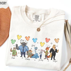 Puede incluir: Camiseta de color marfil con personajes de dibujos animados sosteniendo globos con forma de Mickey Mouse. Los personajes incluyen Flash, Bogo, Nibbles, Gary, Judy Hopps, Nick Wilde y Mayor. La camiseta tiene una etiqueta Comfort Colors y un collar dorado.
