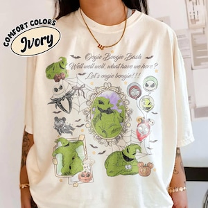 May include: Ivory-colored t-shirt featuring a graphic design with the text "Oogie Boogie Bash" and various characters. The design includes images of Oogie Boogie, Jack Skellington, and other characters from the movie. The shirt has a crew neck and short sleeves.