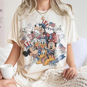 Może przedstawiać: Kremowa koszulka z grafiką postaci Disneya: Myszki Miki, Myszki Minnie, Kaczora Donalda, Kaczki Daisy, Goofy'ego i Plutona, z amerykańskimi flagami i patriotycznymi wzorami. Koszulka zawiera napis "America 250th USA Anniversary 1776-2026".