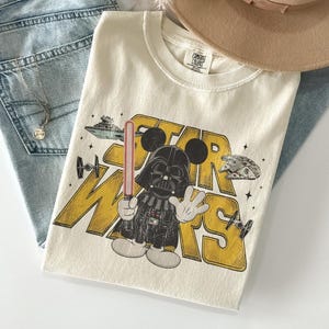 Puede incluir: Camiseta color crema con un gr&aacute;fico de Darth Vader como Mickey Mouse con las palabras "STAR WARS" en amarillo. La camiseta incluye im&aacute;genes de naves espaciales y un sable de luz. La camiseta est&aacute; doblada sobre unos vaqueros azules y un sombrero beige.