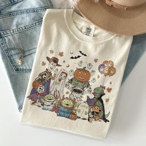 May include: Cream-coloured t-shirt with a Halloween graphic featuring cartoon characters such as Woody and Buzz Lightyear. The design includes pumpkins, ghosts, bats, and autumn leaves. The shirt is folded on a light blue denim surface.