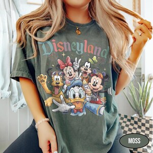 May include: Moss green t-shirt featuring the word "Disneyland" in a colorful, stylized font above a graphic of Disney characters. The graphic includes Mickey Mouse, Minnie Mouse, Donald Duck, Goofy, and Pluto. The shirt is a relaxed fit.
