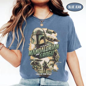 May include: Blue jean-colored t-shirt featuring a graphic from the Star Wars series, "The Mandalorian and Grogu." The design includes images of the Mandalorian, Grogu, and other characters, with the phrase "THIS IS THE WAY" at the bottom.