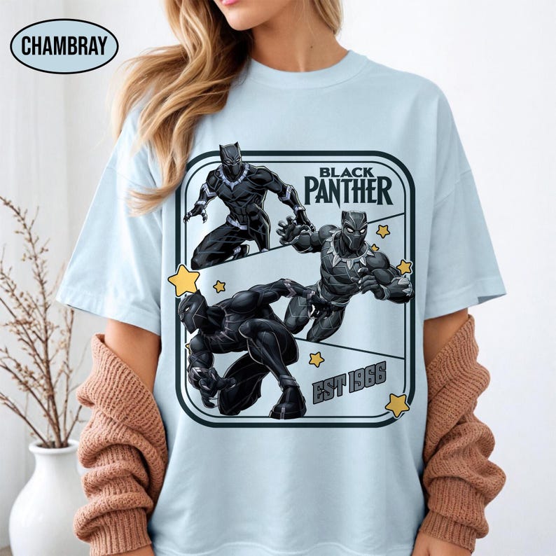 Może przedstawiać: Jasnoniebieska koszulka z grafiką Black Panther w pozach akcji, z tekstem "BLACK PANTHER" i "EST 1966" w prostokątnej ramce. Koszulka ma okrągły dekolt i kr&oacute;tkie rękawy.