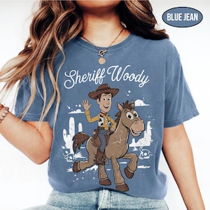 May include: A blue jean colored t-shirt featuring a graphic print of the character Sheriff Woody riding a horse. The design includes the text Sheriff Woody in white script, a desert landscape with cacti, and a skull. The shirt has a crew neckline.