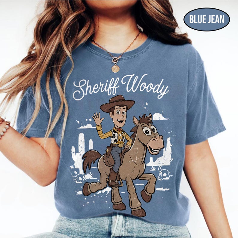 May include: A blue jean colored t-shirt featuring a graphic print of the character Sheriff Woody riding a horse. The design includes the text Sheriff Woody in white script, a desert landscape with cacti, and a skull. The shirt has a crew neckline.