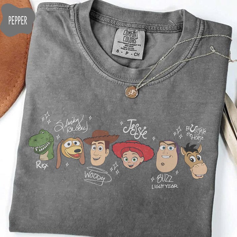 May include: A charcoal gray crew neck t-shirt featuring a graphic print of six animated characters including Rex, Slinky Dog, Woody, Jessie, Buzz Lightyear, and Bullseye. Each character is accompanied by their name written in a white script font.